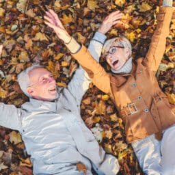 Cheerful active senior couple lying on ground rolling around in autumn leaves having fun