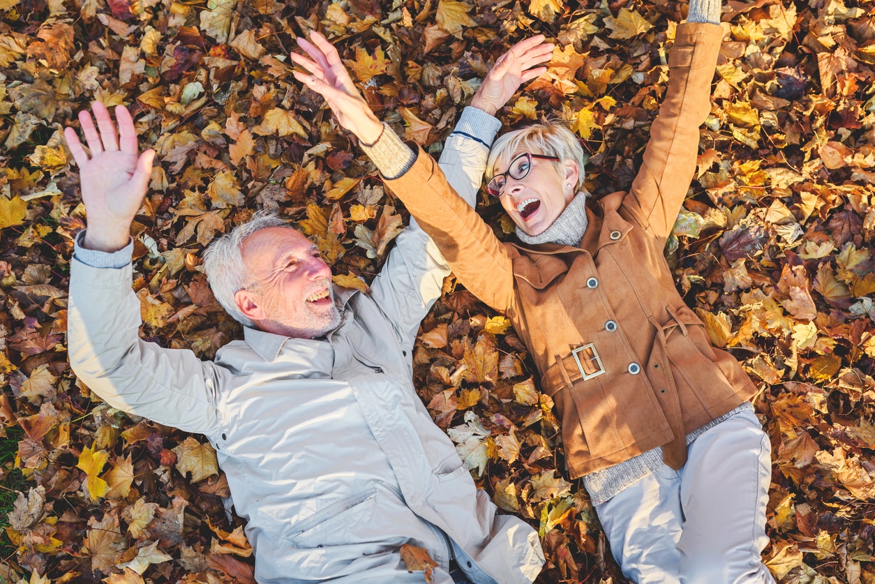 Cheerful active senior couple lying on ground rolling around in autumn leaves having fun