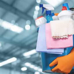 A person in orange gloves carries a bucket of cleaning supplies indoors.