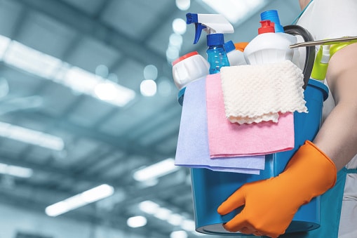 A person in orange gloves carries a bucket of cleaning supplies indoors.