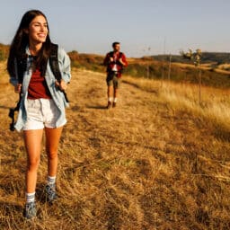 Active young couple having fun hiking in nature, woman carrying backpack walking in foreground, enjoying sunny summer day outdoors