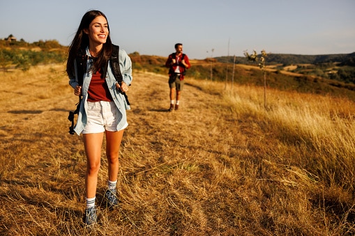 Active young couple having fun hiking in nature, woman carrying backpack walking in foreground, enjoying sunny summer day outdoors