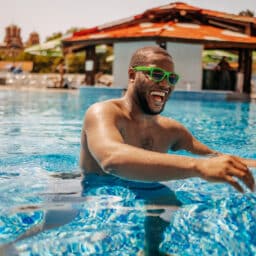 Man enjoying at outdoor swimming pool on sunny day wearing sunglasses while reaching for beach ball