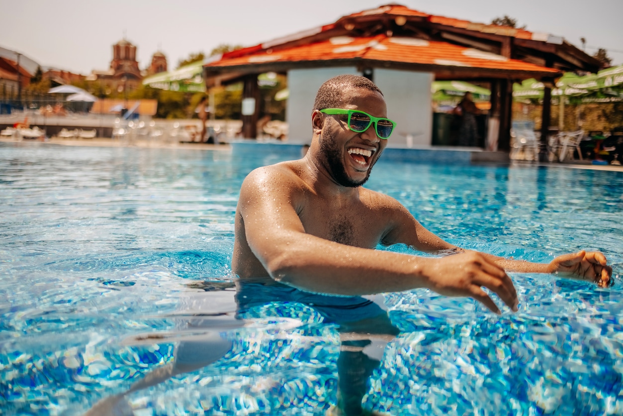 Man enjoying at outdoor swimming pool on sunny day wearing sunglasses while reaching for beach ball.