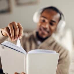 Mid-thirties man with headphones smiling as he reads and turns pages at home, enjoying an audiobook and relaxing in a cozy, modern domestic setting for study and leisure.