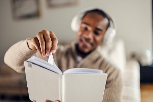 Mid-thirties man with headphones smiling as he reads and turns pages at home, enjoying an audiobook and relaxing in a cozy, modern domestic setting for study and leisure.